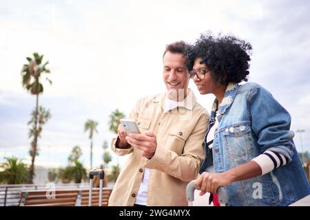 Concentrated multiracial mature tourist couple smiling standing looking and using mobile phone. Stock Photo