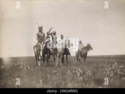 The war party, 1905. Photograph shows a group of Crow Indians on horseback. Chief, in middle, wearing headdress. Stock Photo