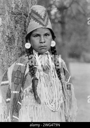 Umatilla maiden, c1910. Umatilla woman, half-length portrait, facing front, standing in front of tree, wearing beaded buckskin dress, shell bead necklaces, shell disk earrings, and woven grass hat. Stock Photo