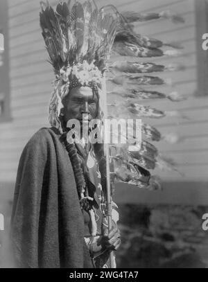 Piegan war-bonnet and coup-stick, c1910. Piegan man, half-length portrait, dressed in war bonnet and holding a feathered coup-stick, facing front. Stock Photo