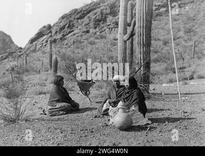 Qahatika women resting in Harvest Field-Qahatika, c1907. Three Qahatika women sitting on ground with kiho carriers and pot nearby, saguaro cacti and mountains in background. Stock Photo