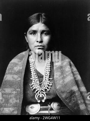 Head-and-shoulders portrait of Navajo woman, facing front, c1904. Navajo woman wearing silver squash blossom necklace, concho belt, blanket draped over shoulders. Stock Photo