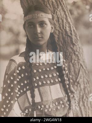 Lucille [B], c1907. Photograph shows Lucille, a Native woman, half-length portrait, standing next to tree, facing front, wearing a headband and seashell decorated buckskin dress. Stock Photo