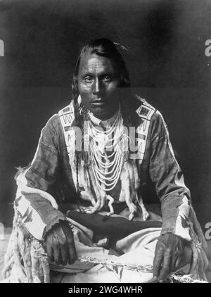 Wades in Water, Piegan Indian, full-length portrait, seated on floor, facing front, with braids, beaded buckskin shirt and leggings, beads with ermine tail trim, c1910. Stock Photo