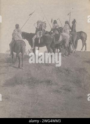 Overlooking the Little Horn, 1908. Photograph shows five Crow Indians, Wolf, Fish Shows (wearing headdress), Which Way, Lone Tree, and Two Whistles on horseback carrying staffs. Stock Photo