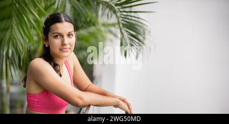 Relaxed young woman in a pink sports bra resting elbows on a balcony railing Stock Photo
