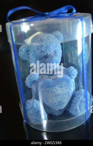 The blue teddy bear made of natural wax is made in the shape of a candle and packed in a plastic box with a blue bow on a black glossy background. Stock Photo