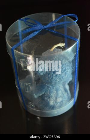 The blue teddy bear made of natural wax is made in the shape of a candle and packed in a plastic box with a blue bow on a black glossy background. Stock Photo