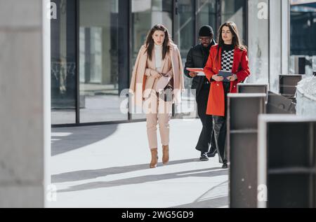Two businesswomen and a businessman engrossed in conversation while walking through a modern city environment on a sunny day. Stock Photo