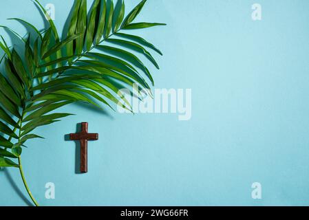 Palm Sunday. Wooden christian cross and palm leaves on blue background, top view, copy space. Palm sunday holiday concept. Stock Photo