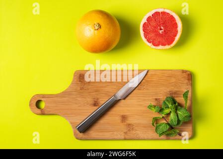 Cutting board with a knife and mint, next to a red grapefruit cut in half and another whole, greenish background. Stock Photo