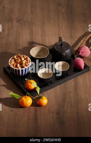 A black tea set with cashew nuts, mandarin oranges and pink flowers on wooden background. Holiday content Stock Photo
