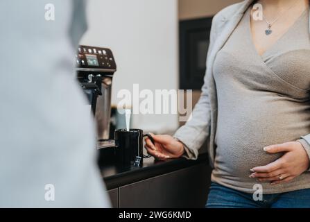 Pregnant businesswoman enjoying her tea during break at work. She is standing at the office kitchen, discussing work with her colleague. Stock Photo