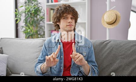 A young hispanic man with a beard and denim jacket sits in a modern living room making hand signs. Stock Photo