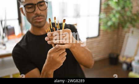 A focused young hispanic man examines paintbrushes in a creative studio setting. Stock Photo