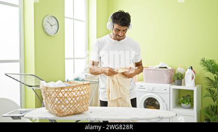 A young hispanic man folding laundry in a bright green room with a washing machine, headphones, ironing board, and laundry basket. Stock Photo