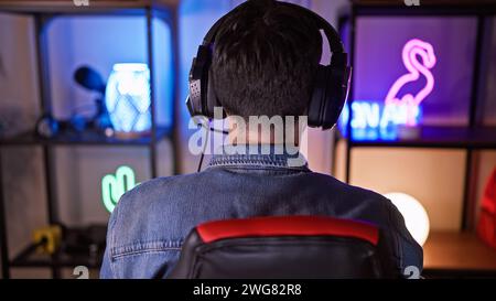 Rear view of a young hispanic man in a gaming room wearing headphones and a denim jacket at night Stock Photo