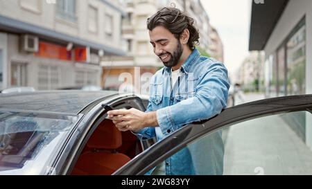 Young hispanic man using smartphone leaning by door car at street Stock Photo