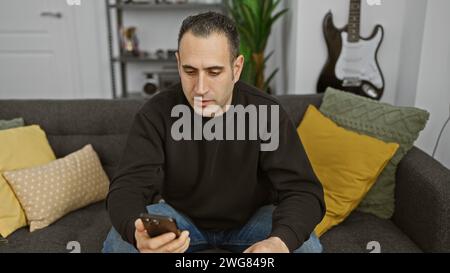 A hispanic man casually dressed in a sweater and jeans using a smartphone while seated on a grey sofa with colorful pillows indoors. Stock Photo