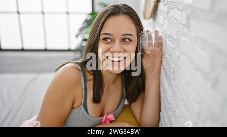 Intriguingly nosy young hispanic woman with attractive hairstyle, eavesdropping through bedroom wall with a glass, curious about private neighbour gos Stock Photo