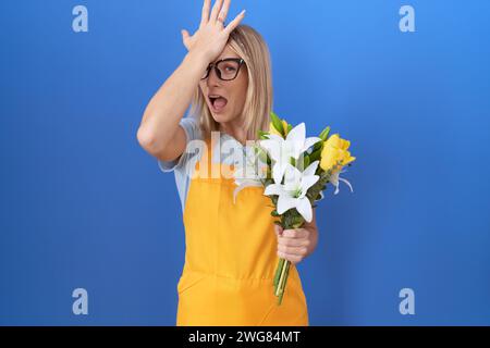 Young caucasian woman wearing florist apron holding flowers surprised with hand on head for mistake, remember error. forgot, bad memory concept. Stock Photo