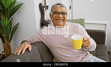 A handsome, middle-aged man with grey hair relaxes in a modern living room, smiling and holding a yellow mug. Stock Photo
