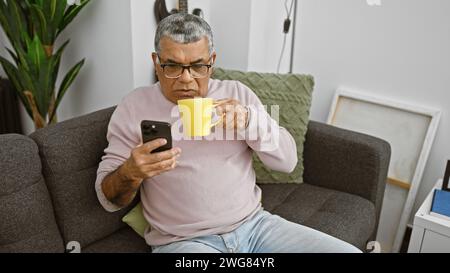 Middle-aged man with glasses using smartphone and holding mug in the living room Stock Photo