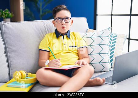 Young hispanic kid doing homework sitting on the sofa making fish face with lips, crazy and comical gesture. funny expression. Stock Photo