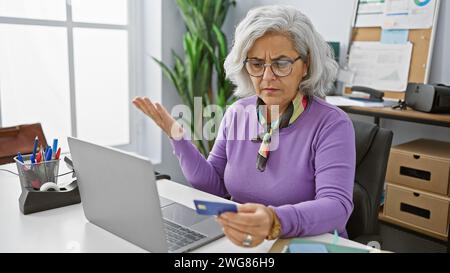Confused elderly woman holding credit card in modern office looking perplexed at laptop screen. Stock Photo