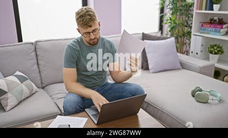 Handsome caucasian man with blue eyes and a beard reviews documents on a laptop in a modern living room Stock Photo