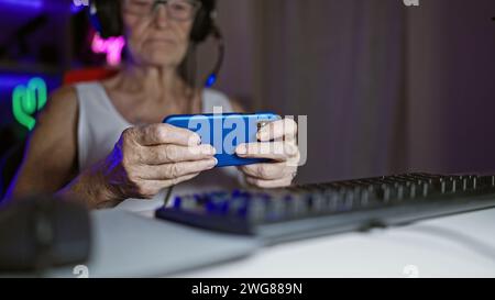 Cool grey-haired senior woman streamer rocks the gaming world, playing video game on smartphone, immersed in virtual entertainment, middle-aged but di Stock Photo