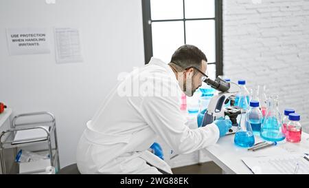 Hispanic scientist man analyzing samples with a microscope in a laboratory setting wearing lab coat and gloves. Stock Photo