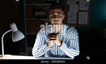 A contemplative young hispanic man sits indoors at a dimly lit office desk, emanating an air of focused seriousness. Stock Photo