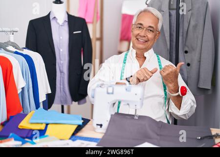 Middle age man with grey hair dressmaker using sewing machine pointing to the back behind with hand and thumbs up, smiling confident Stock Photo