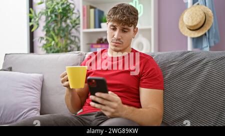 Handsome young hispanic man relaxing at home, concentrated on texting while drinking coffee on sofa Stock Photo