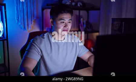 A young asian man focused while using a computer in a dark room with ambient lighting. Stock Photo