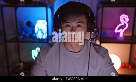 A young asian man wearing headphones sits engaged in his neon-lit gaming room at home during the night. Stock Photo