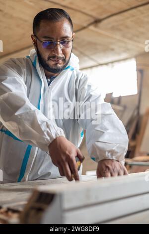 workplace with young man with glasses and overalls measuring to perform cutting, factory with furniture machinery, lifestyle and tools Stock Photo