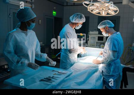 Remote view of skilled diverse surgeons and nurse wearing sterile gloves, surgical uniforms and masks processing surgical operation in dark operating Stock Photo