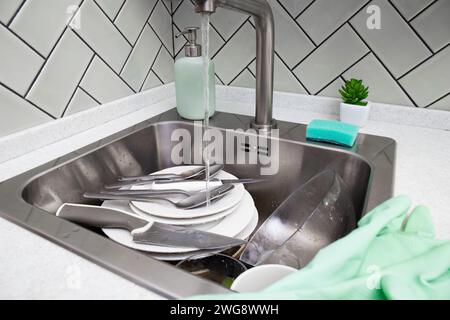 A stainless steel sink contains a stack of used plates and utensils, with running water, soap dispenser, and cleaning sponge ready for the task of dis Stock Photo