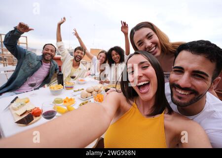 Excited young pretty woman taking group selfie with joyful friends on rooftop. Cheerful people photo Stock Photo