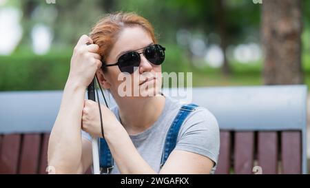 Blind red-haired woman sitting outdoors leaning on a cane.  Stock Photo