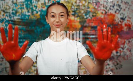 Happy highschool girl looking at camera while showing two side of hand to camera in art lesson. Smiling caucasian student wearing white shirt while Stock Photo