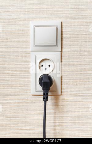 A beige double socket with a black electric plug and a single switch on a light wall. Stock Photo