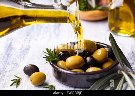 in the foreground, extra virgin olive oil is poured into a bowl with rosemary-flavored olives Stock Photo