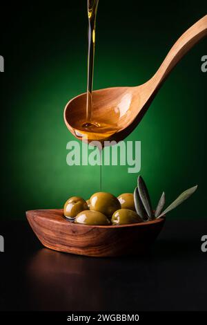 from the wooden spoon, in the foreground, the extra virgin olive oil is poured into an olive wood bowl with olives Stock Photo