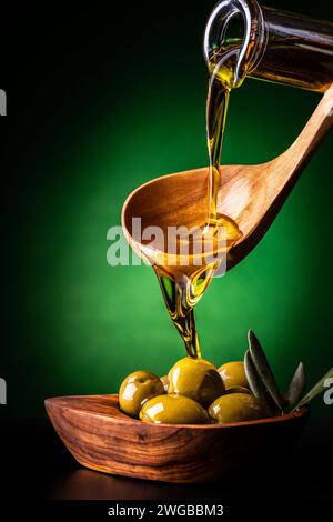 from the wooden spoon, in the foreground, the extra virgin olive oil is poured into an olive wood bowl with olives Stock Photo