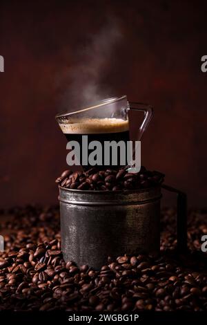an old metal measuring cup filled with roasted coffee beans, on top of a steaming cup of espresso. Stock Photo