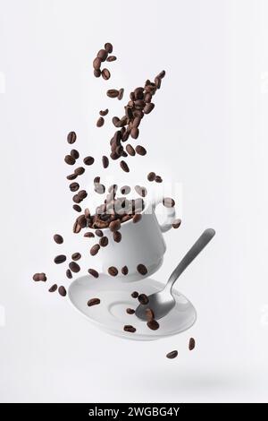 roasted coffee beans spilled from a floating cup and a teaspoon isolated on white background Stock Photo