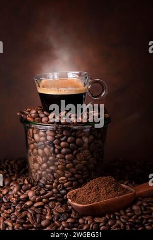 a glass jar filled with roasted coffee beans, on top of a steaming cup of espresso. Stock Photo